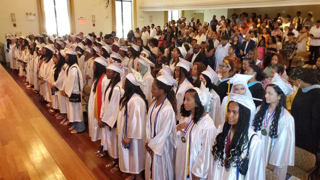 High school students in graduation caps and gown stand in auditorium, shared in newsletter of school year updates