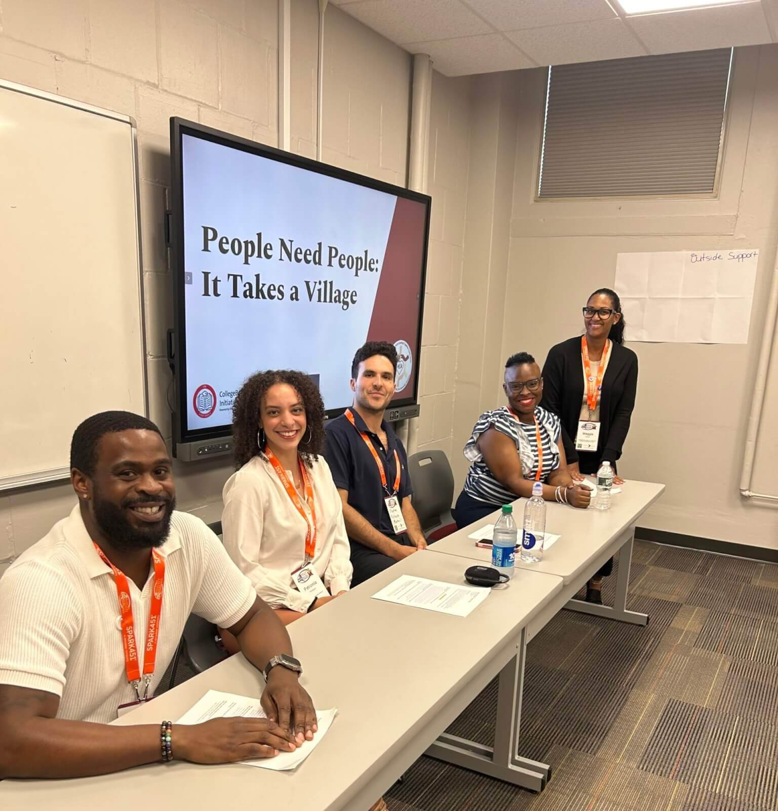 Five adult presenters sit behind a table, with a screen behind them titled "People Need People: It Takes a Village" at the NYSACAC conference
