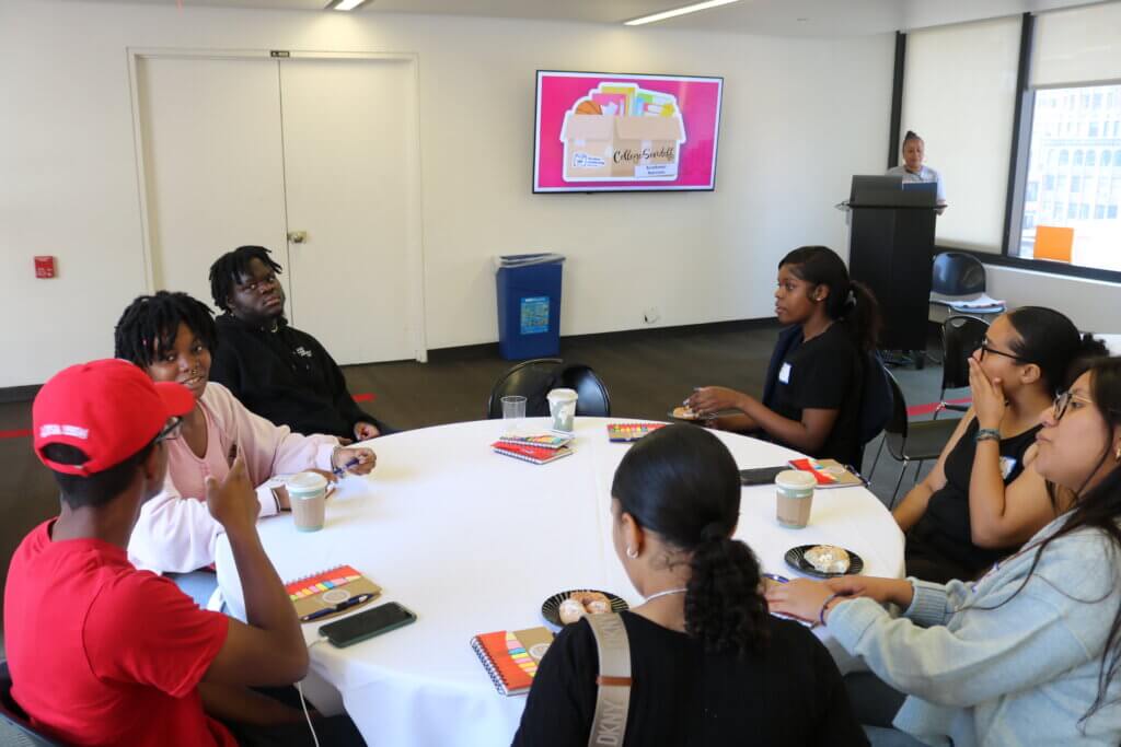 Seven students sit around a table, in conversation with each other