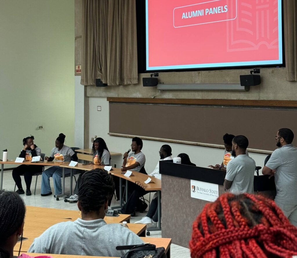 High school alumni are seated in front of lecture hall, with large red screen behind them with text "Alumni Panels"