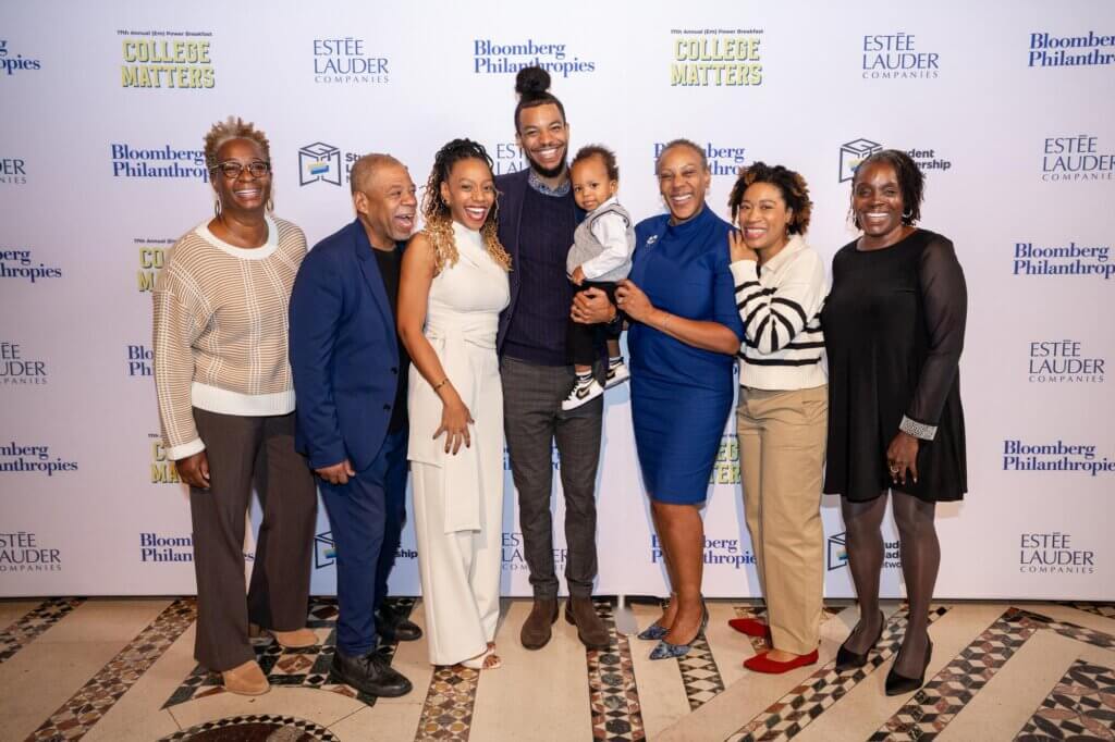 Family and guests, including a smiling toddler, in professional and festive attire pose for photo in front of event backdrop
