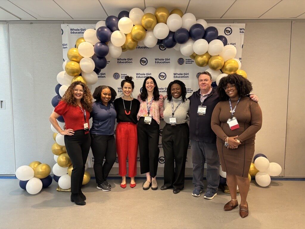members of the Girls' Education team in professional wear pose for photo in front of backdrop with Student Leadership Network logos