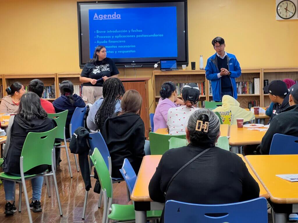 Two educators stand in front of screen with Spanish text about the college process, while high school students are seated