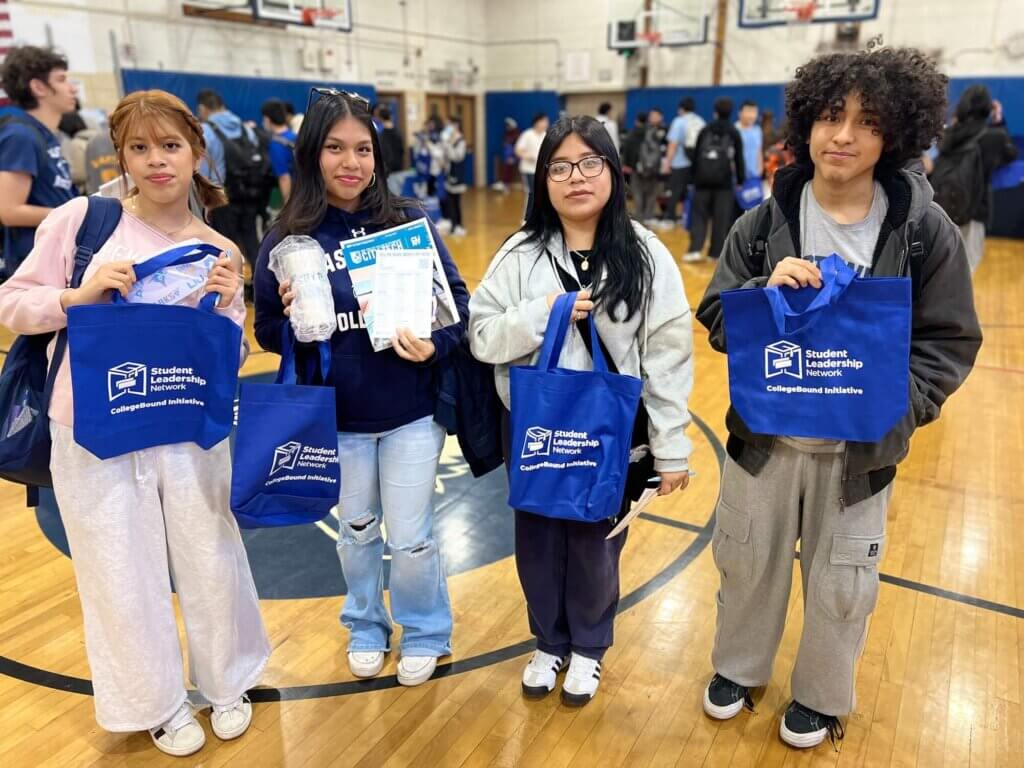 Four students hold up Student Leadership Network bags and brochures while navigating college fairs at school