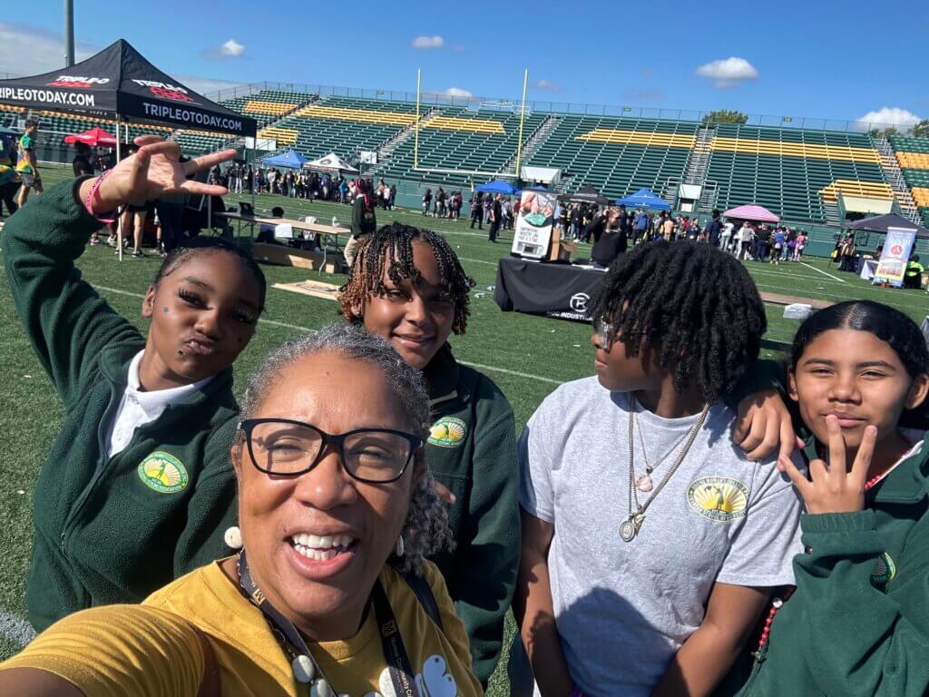 College counselor takes selfie with four female students on a football field with college tables