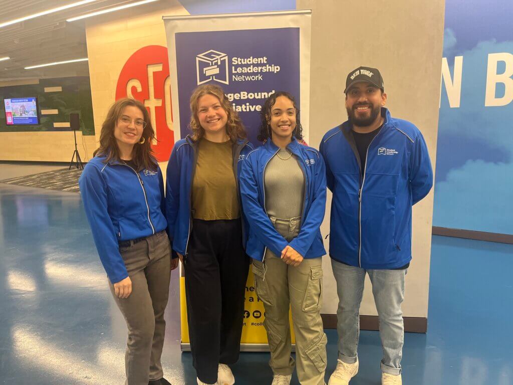 Four college counselors with matching blue staff jackets stand in front of Student Leadership Network sign