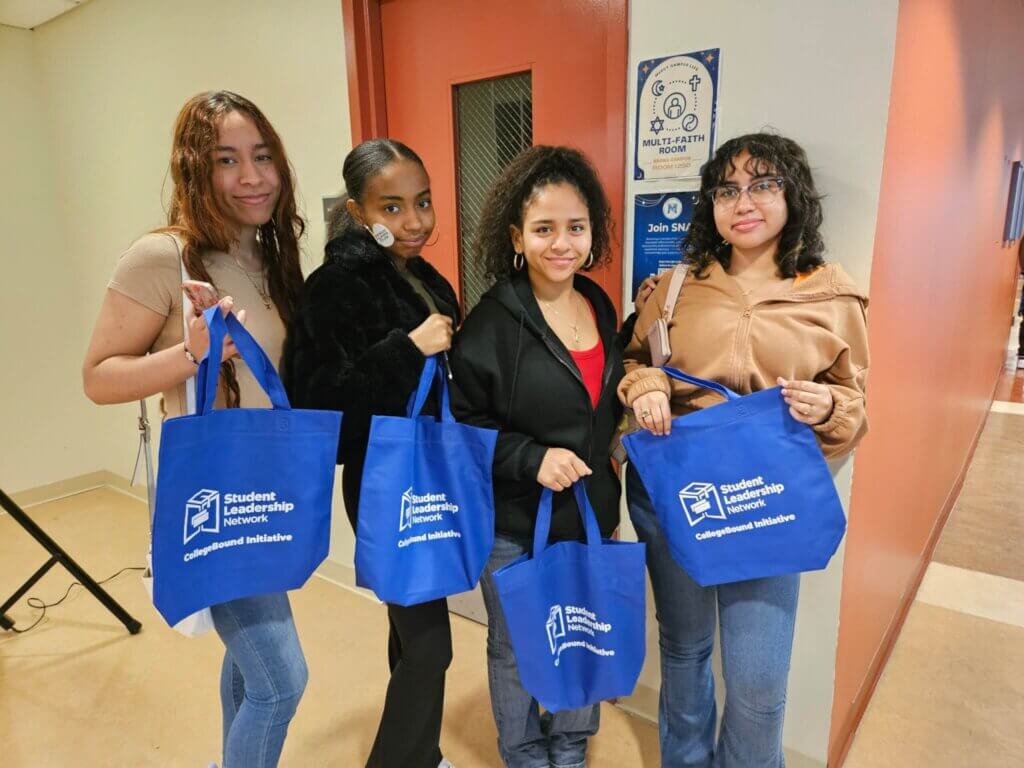 Four young women stand indoors holding blue tote bags labeled Student Leadership Network: CollegeBound Initiative, showcasing their participation at the Bronx Collaborative College Fair 2024