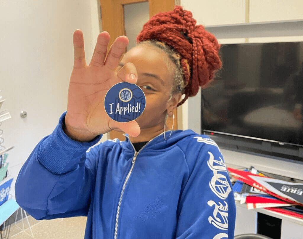 A person in a blue jacket holds up a round I Applied! badge in front of their face, standing in their director of college counseling's office, showcasing their involvement with the Student Leadership Network.