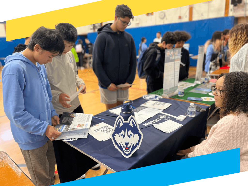 Students gather around a table with a UConn husky logo, reading pamphlets and discussing opportunities with a representative at an event inspired by the NCAN Conference in the gymnasium.