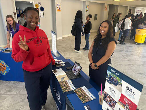 Two women stand at an NCAN Conference college fair table with brochures and a tablet; one smiles at the camera while the other flashes a peace sign. Other attendees can be seen in the background.