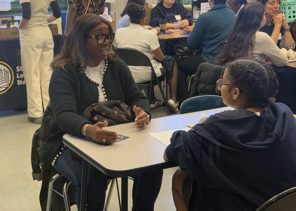 Volunteer is seated at desk talking with a student