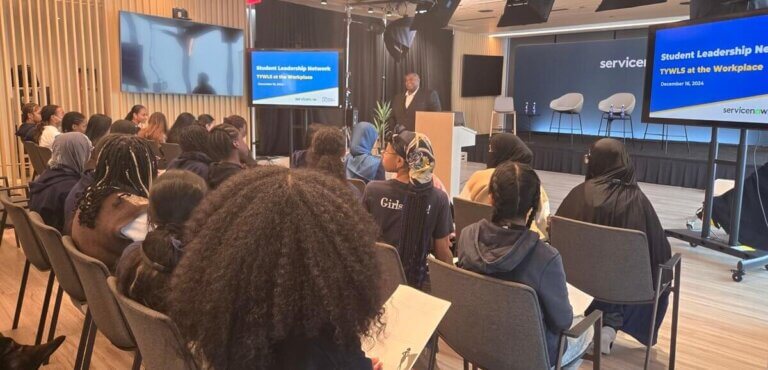 A man is smiling at the front of the room, while greeting students who are seated in the audience