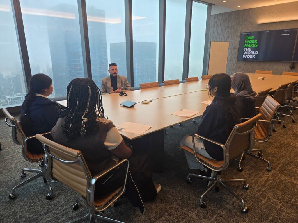 Adult volunteer and four students are talking with each other while seated at a conference table