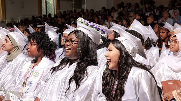 High school girls+ wearing graduation caps and gowns are seated in an auditorium