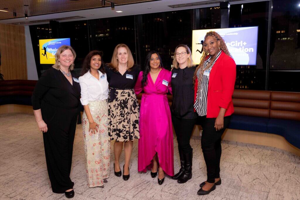 Six women stand together and smile for a group photo at the WholeGirl+ Education National Conference, with name tags visible and a presentation screen in the background.