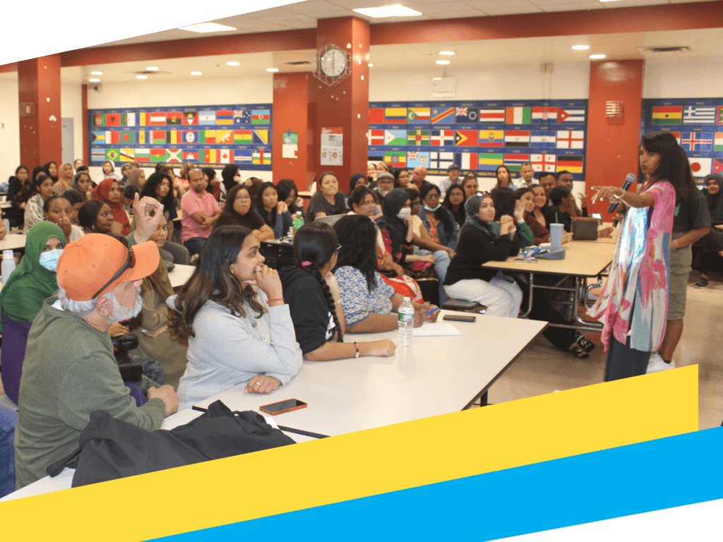 A woman stands and speaks to a large seated group in a cafeteria decorated with international flags, sharing insights about college access.