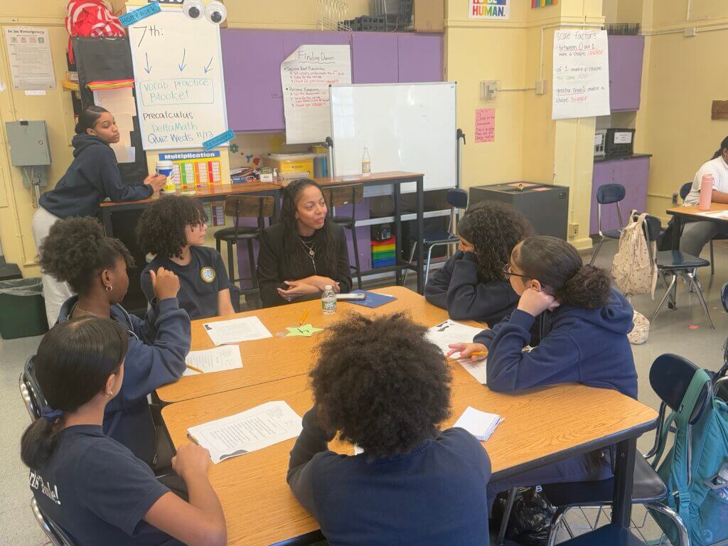 adult sits in round table with six students who are asking her questions