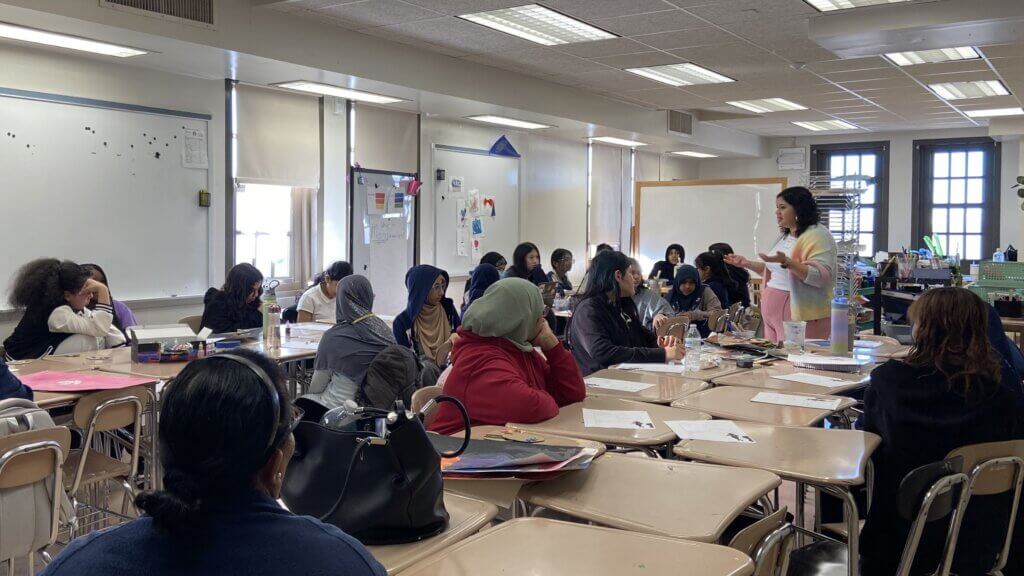 adult woman standing with arms gesturing, speaking to a classroom of female students