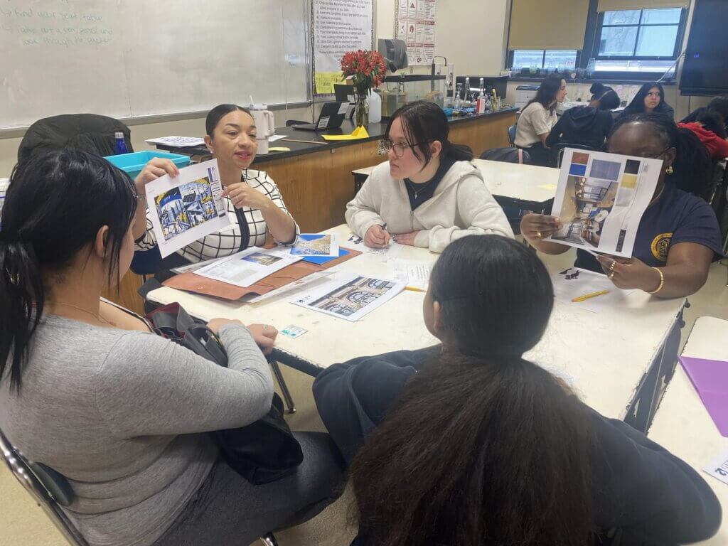 adult volunteer holds up a printout of real estate designs to a table of female students