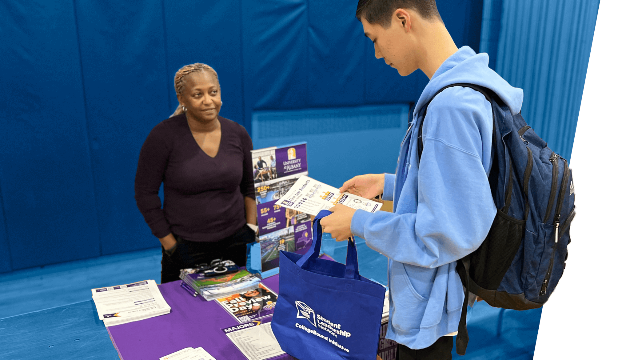 A student holding flyers stands at a table with informational materials about equitable college counseling, speaking to a woman at an event or fair.