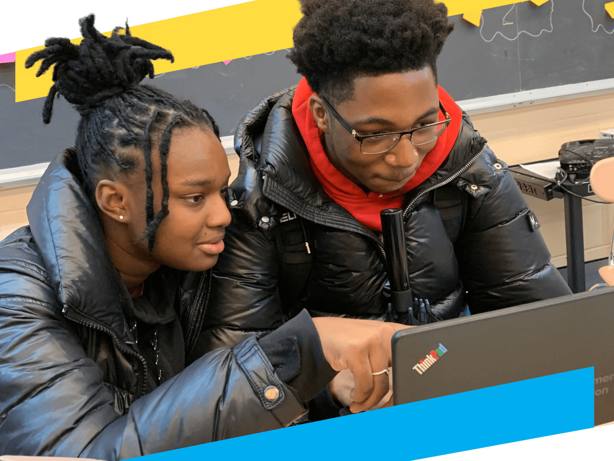 Two students sit at a desk in a classroom, pointing to a laptop with an NYC Department of Education sticker. The chalkboard behind shows assignments, as they discuss Student Leadership Network alumni services.