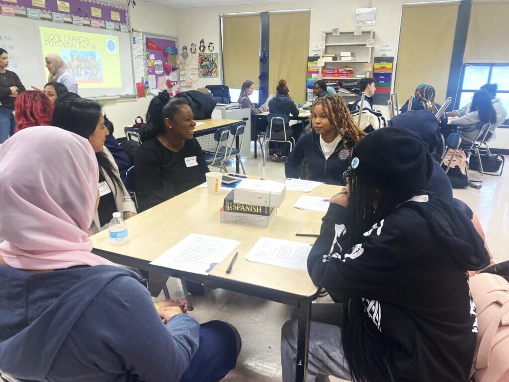 Adult volunteer is seated with students at desks talking