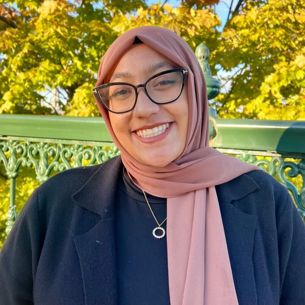 Young woman is smiling confidently, and is wearing glasses and a pink headscarf
