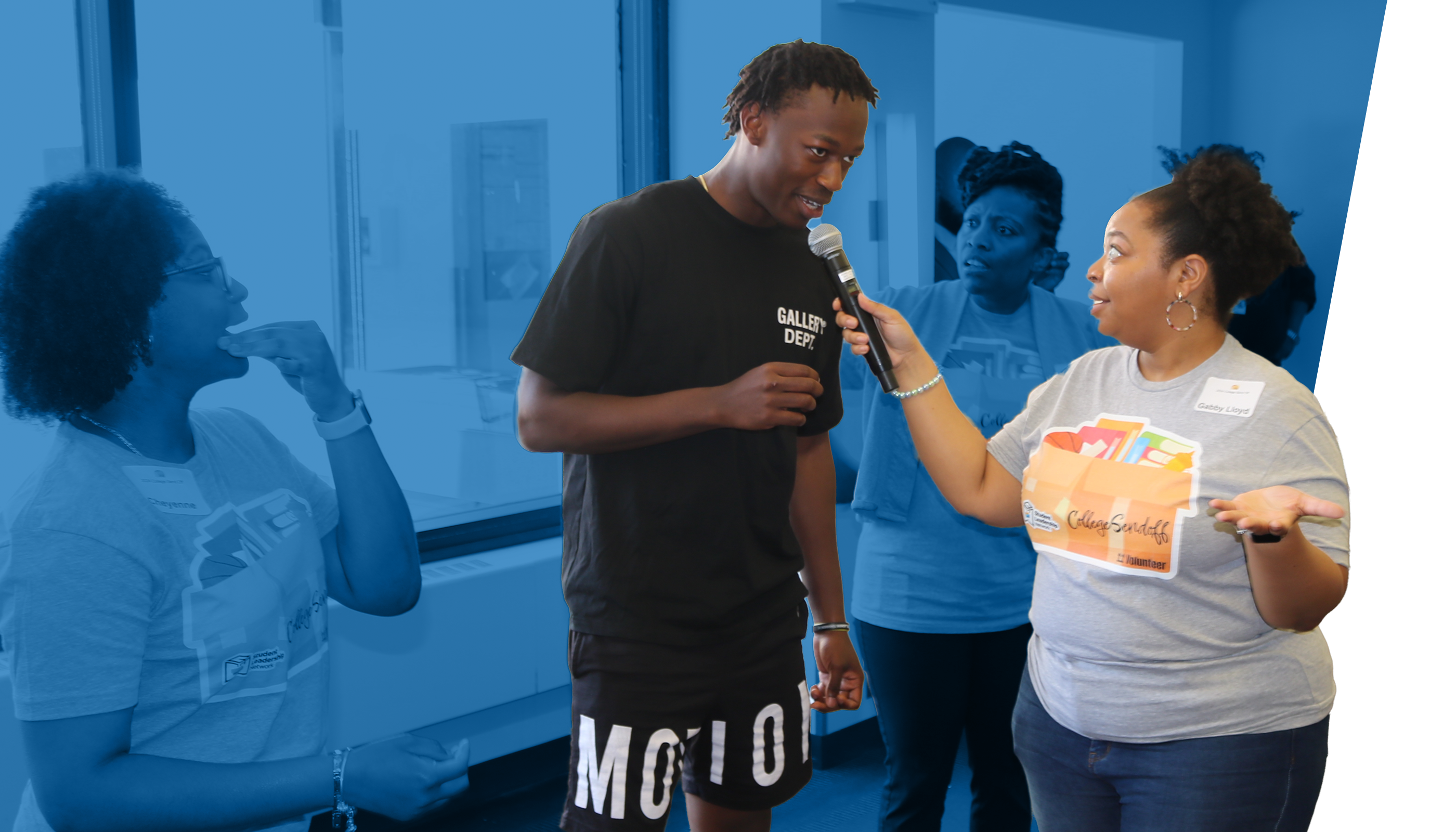 A woman holds a microphone and interviews a young man standing in front of a group in a brightly lit room. Others watch and gesture in the background.