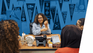 A woman sits at a desk talking to two others, with various college pennants displayed on the wall behind her, reflecting the student leadership networks mission to guide young people toward higher education.