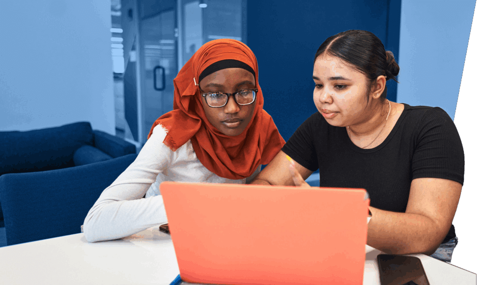 Two young women, both CollegeJourney peer leaders, sit at a table focused on a coral-colored laptop. One wears a red hijab and glasses, while the other wears a black top. A smartphone rests nearby on the table.