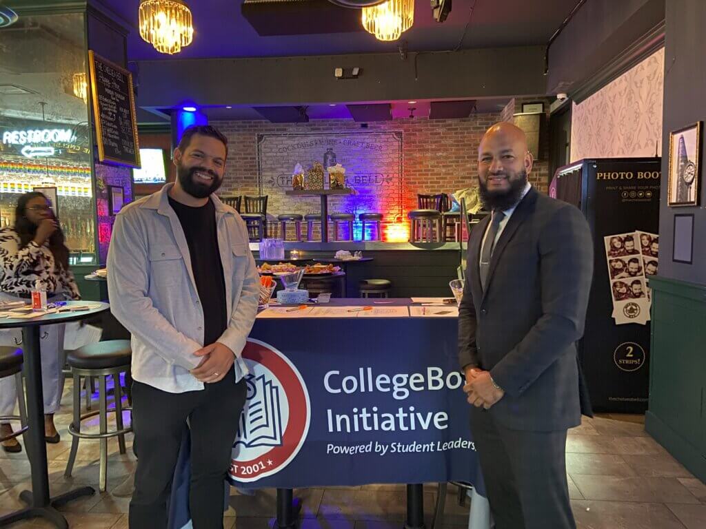 Two men stand in front of a table with a “CollegeBound Initiative” banner, representing Student Leadership Network alumni services at an indoor event venue.