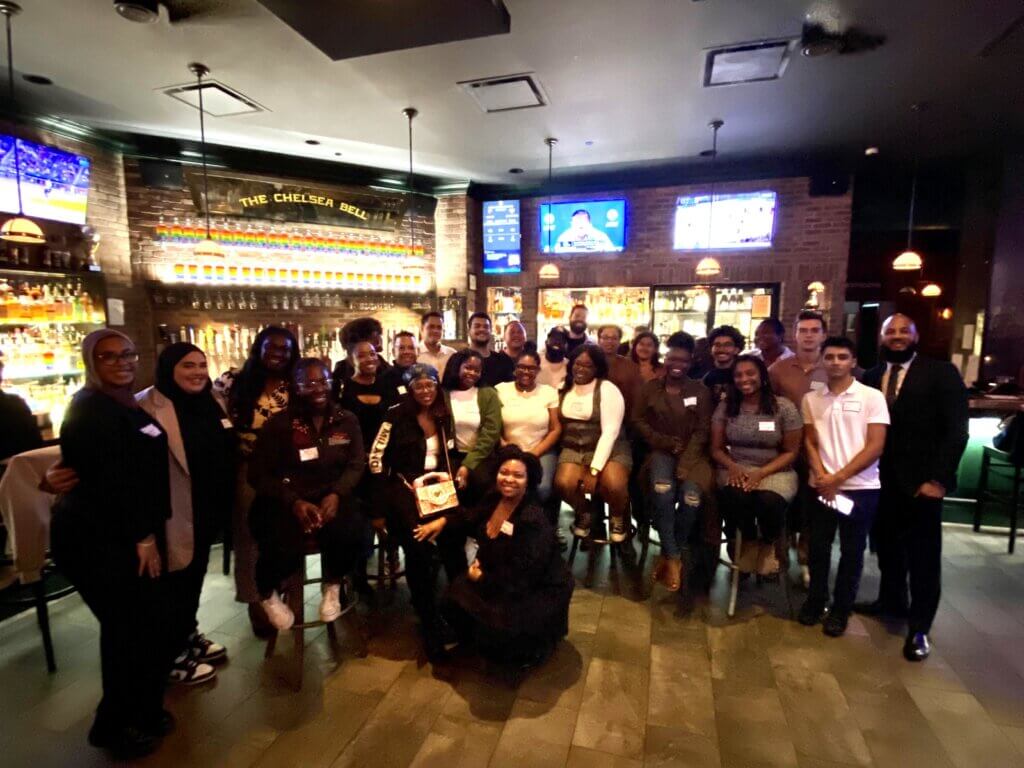 A group of people poses together inside a bar or restaurant, with shelves of bottles, bar stools, and TV screens visible in the background, celebrating Student Leadership Network alumni services.