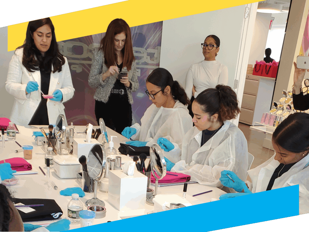 A group of TYWLS at the Workplace volunteers from Intercos, some wearing lab coats and blue gloves, lead a hands-on STEM activity at a table with cosmetic tools. 