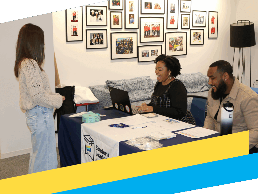 Two people sit at a table assisting a woman standing in front of them; the table has forms, tickets, and a laptop, with framed photos on the wall in the background.