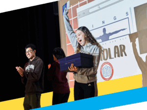 A young woman stands on stage holding a box and an envelope, raising her arm in celebration, while two others applaud beside her—a proud moment in front of a projected presentation.