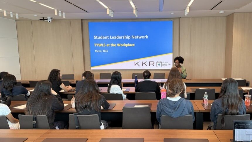 Kristen Gordon speaks into microphone in front of boardroom with students seated in front