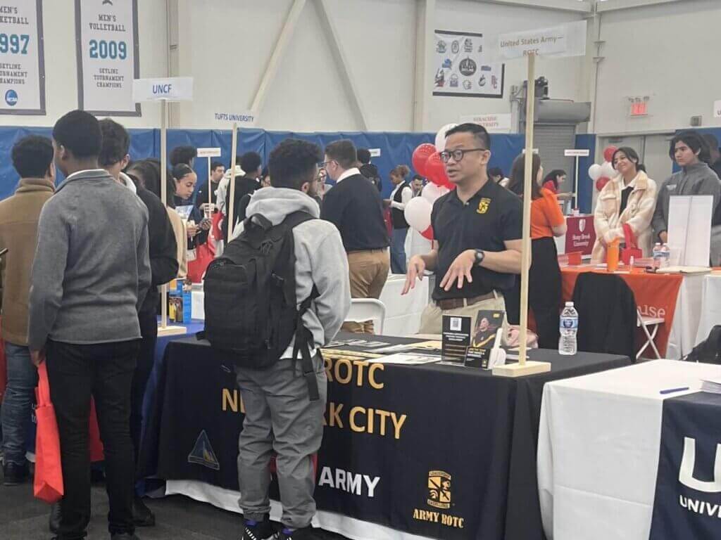 A representative at a New York City Army ROTC booth speaks with students at an indoor college fair during volunteer appreciation week, with other university tables and attendees in the background.