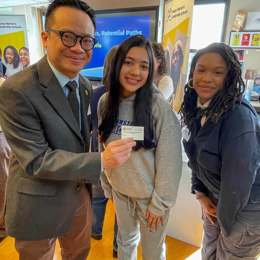 Three people stand indoors; one man in a suit holds an ID card with a young woman in casual clothes, while another woman in a dark outfit smiles beside them during volunteer appreciation week.