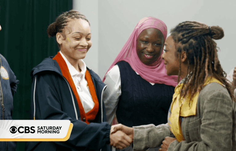 Three women stand indoors, two shaking hands and smiling as a third woman in a pink hijab observes