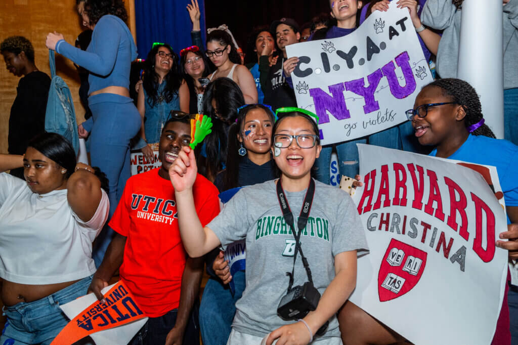 A group of students at an indoor event hold university signs and wear college shirts, smiling and cheering.