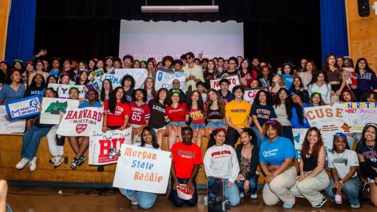 Large group of students posing on a stage, holding college signs and wearing shirts from various universities and colleges, celebrating college decision day