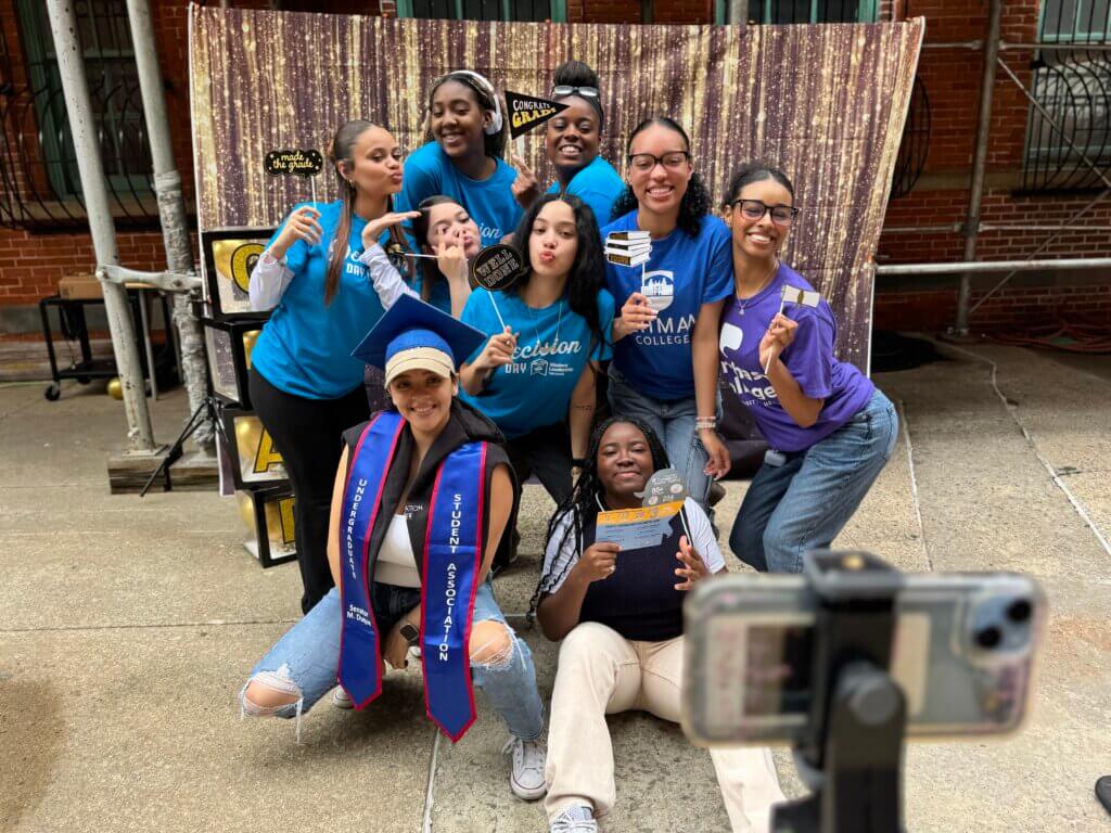 A group of nine young adults pose and smile for a selfie at an outdoor event, some holding props and wearing college-themed shirts, with a sparkling backdrop behind them.