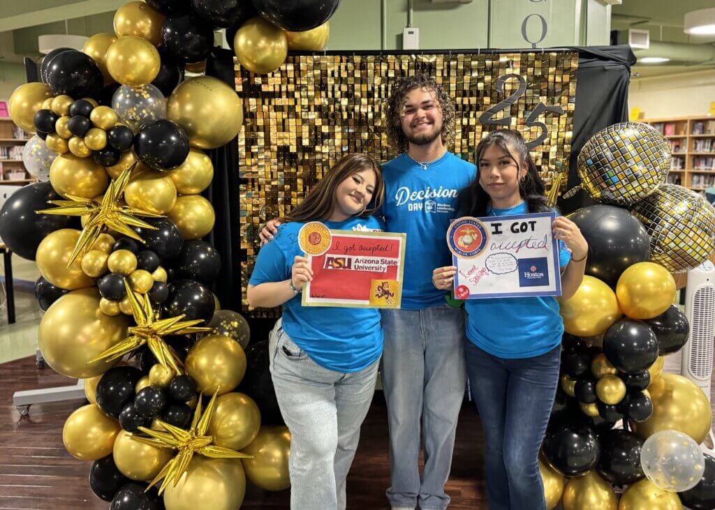 Three students in blue shirts stand in front of a gold and black balloon backdrop, holding college acceptance signs for Arizona State University and Boston University.