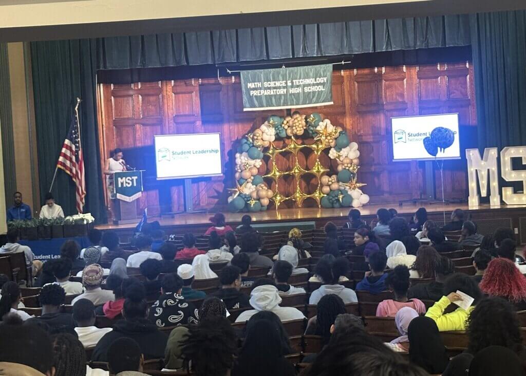 A large group of students sit in an auditorium facing a stage with a Math Science & Technology Preparatory High School banner, balloons, and a speaker at a podium.