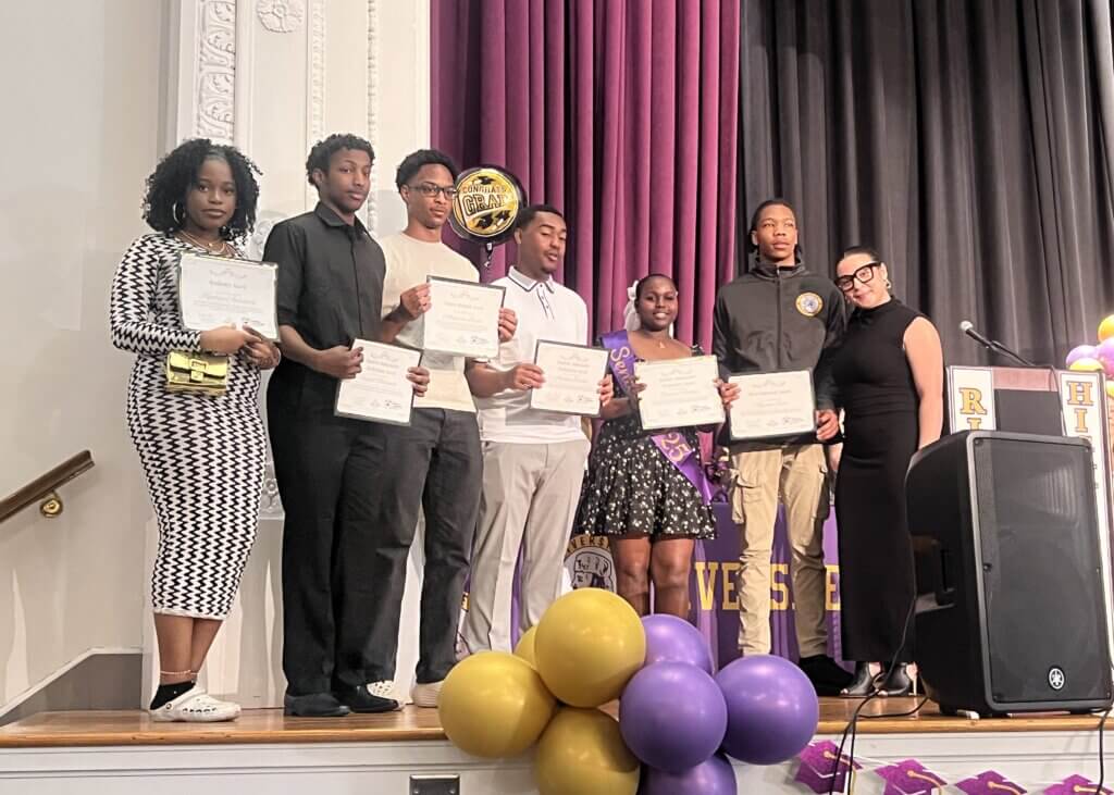 Seven people stand on a stage holding certificates, with purple and gold balloons in front and a podium to the side.
