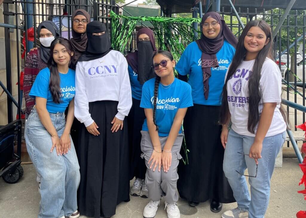 A group of eight young people pose together outdoors, wearing CCNY and Decision Day shirts in front of a green and silver fringe backdrop.
