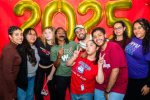 A group of students poses in front of large gold “2025” balloons and a red background, smiling and making peace signs.