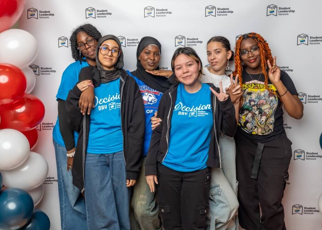 Six young people stand together smiling in front of a Student Leadership Network backdrop, some wearing blue “Decision Day” shirts and casual clothing, with balloons to the side.