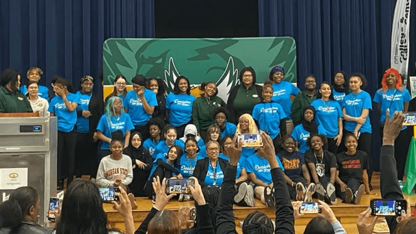 A group of students and adults pose for a group photo on a stage, with several people in the audience photographing them. A green and white logo is displayed in the background.