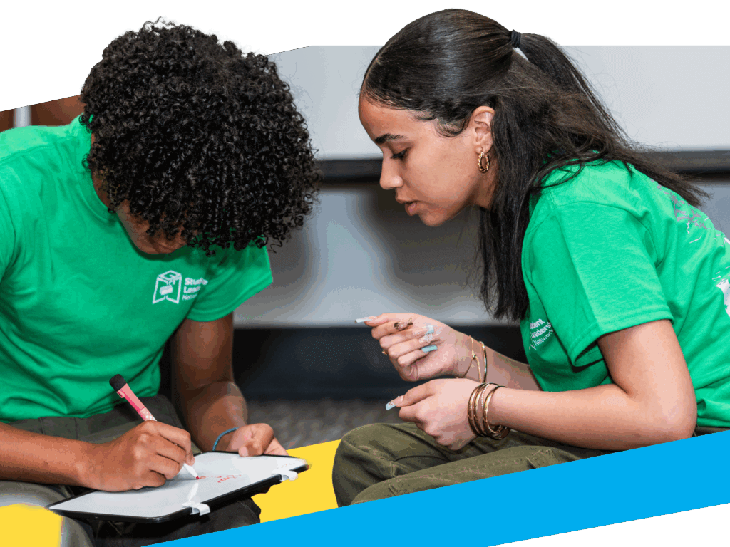 Two students in green shirts sit together; one writes on a small whiteboard while the other watches attentively, both focused on the task—a scene reflecting collegebridge support for transition from high school to college.